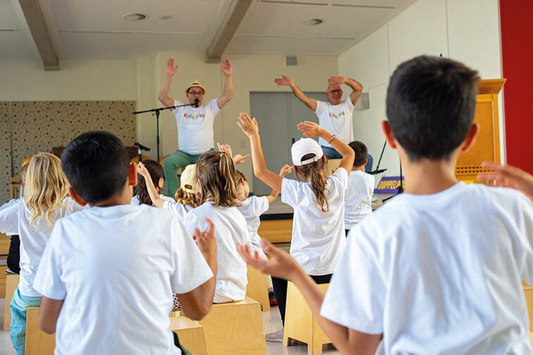 In der Aula zeigen zwei Erwachsene die Choreographie zur Musik, die Kinder tanzen mit.
