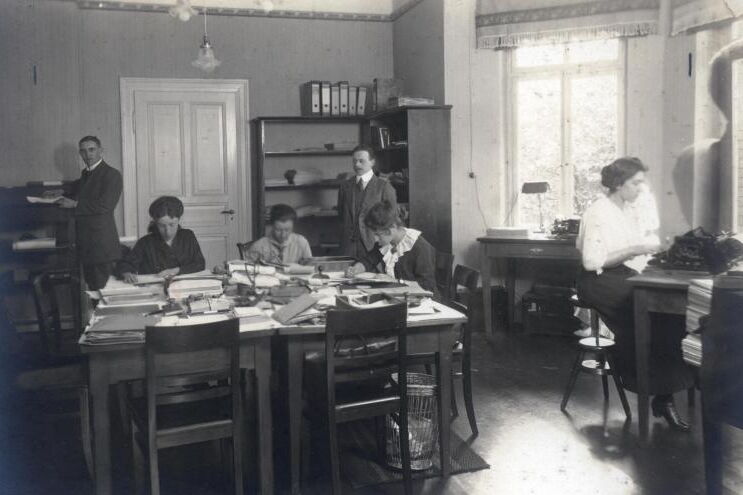 Historische Schwarzweißfotografie: Vier Frauen arbeiten sitzend an Papieren, zwei Männer stehen im Hintergrund.