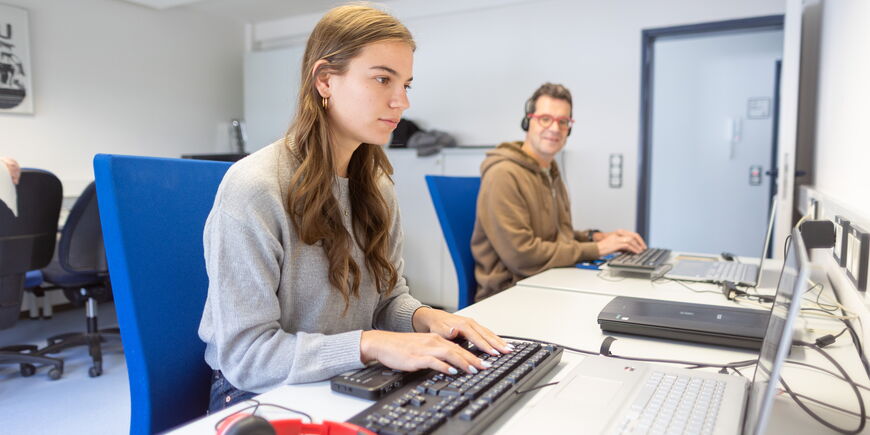 Zwei Teilnehmende sitzen in einem Unterrichtsraum und arbeiten an Laptops.