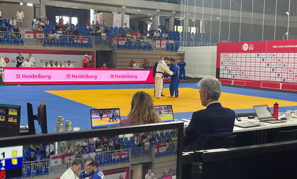 Blick über die Schulter der Juror*innen auf Bildschirme bei der Para-Judo-Europameisterschaft.  Zwei Judoka kämpfen.