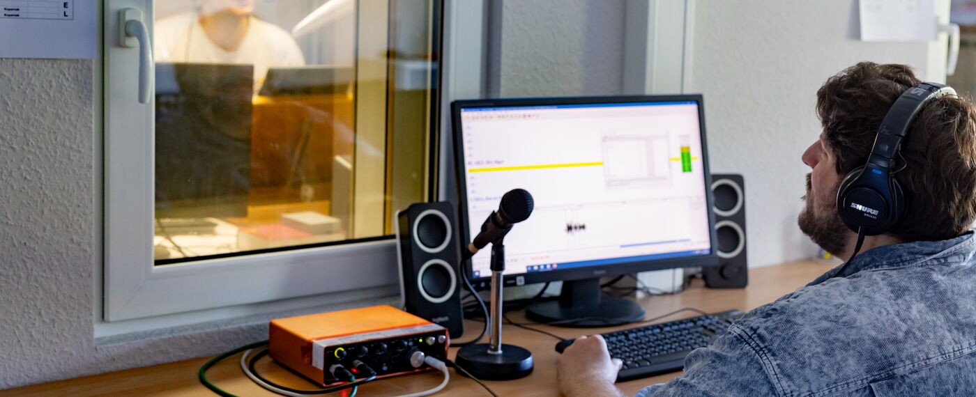 Ein Hörbuchsprecher hinter einem Fenster einer Sprecherkabine, ein Kollege mit Headset außerhalb spricht mit ihm.