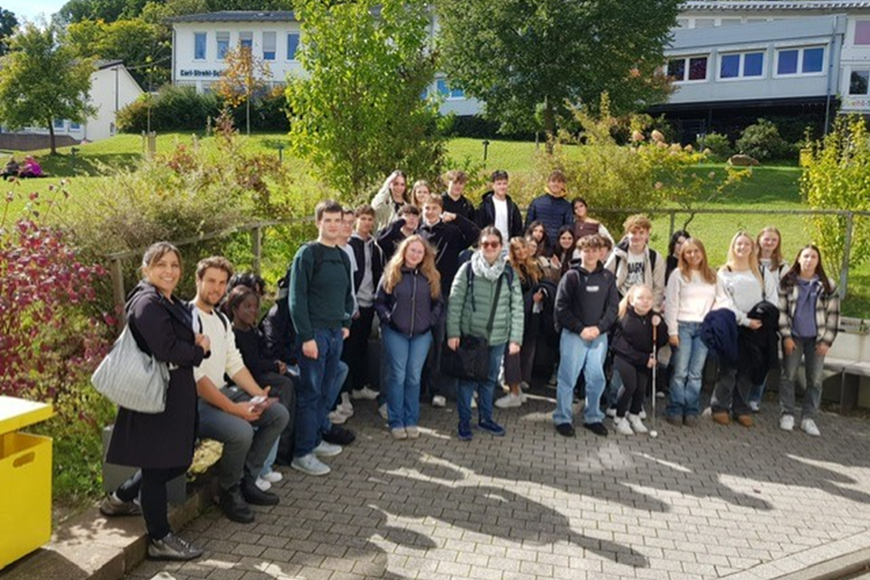 Die gruppe aus blista-Schüler*innen und spanischen Besucher*innen, ca. 30 Personen sitzen und stehen auf und vor einer Bank auf dem blistaCampus.